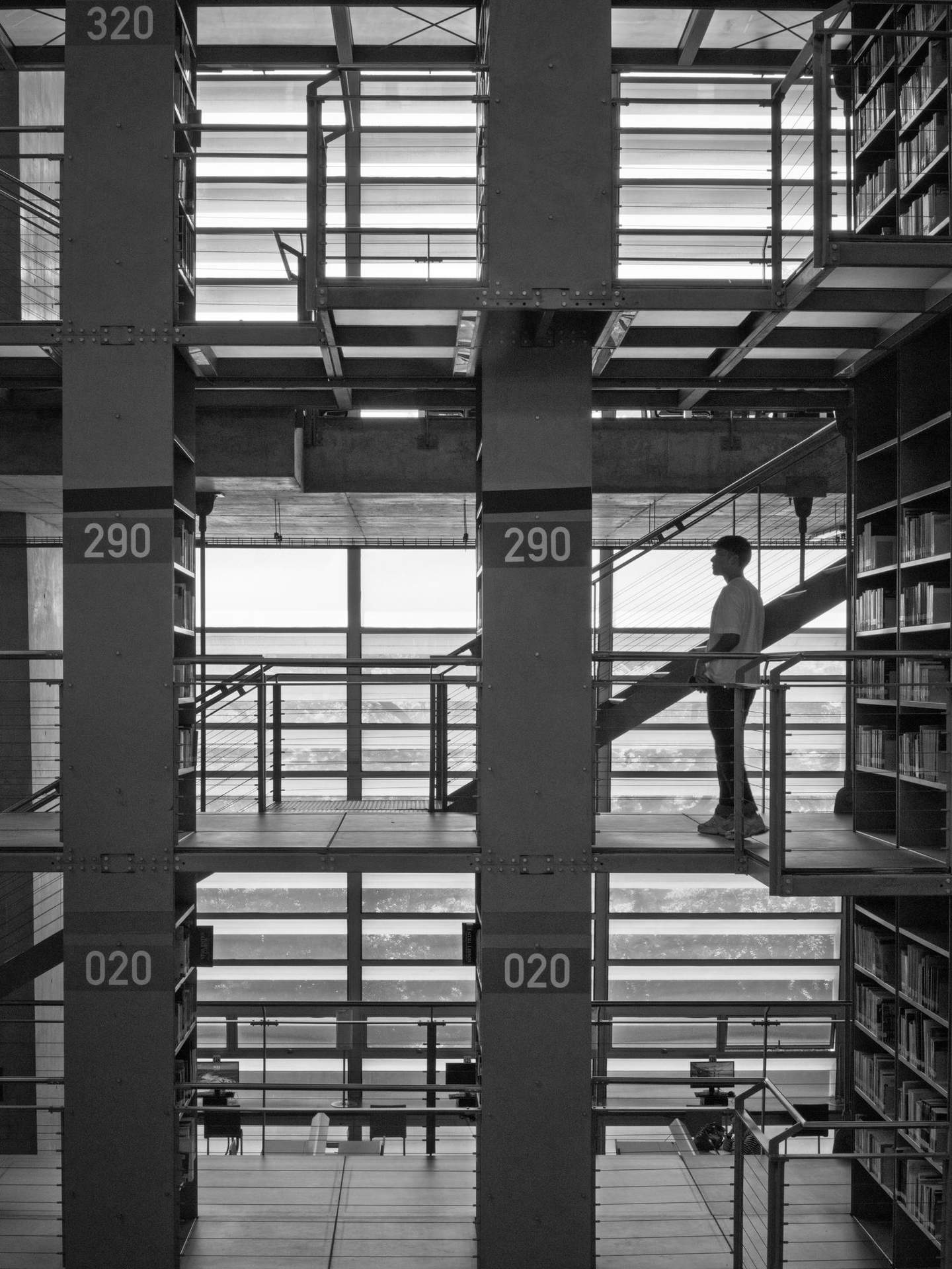 Silhouetted visitor pausing on cantilevered walkway at Biblioteca Vasconcelos, backlit against exterior glazing, black and white