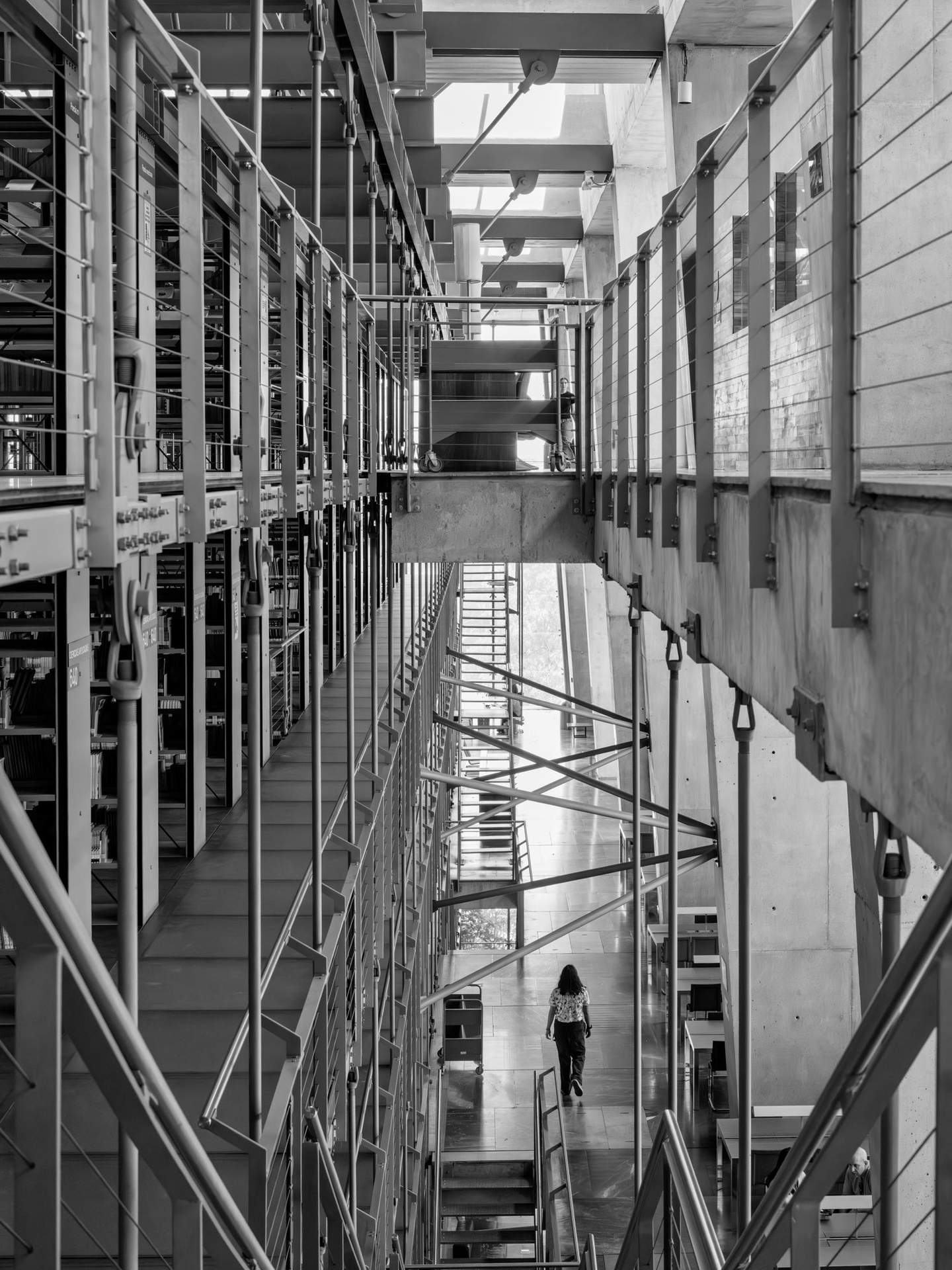 Biblioteca Vasconcelos perimeter circulation corridor in black and white, visitor walking away backlit by exterior glazing, suspended stack structure visible to left
