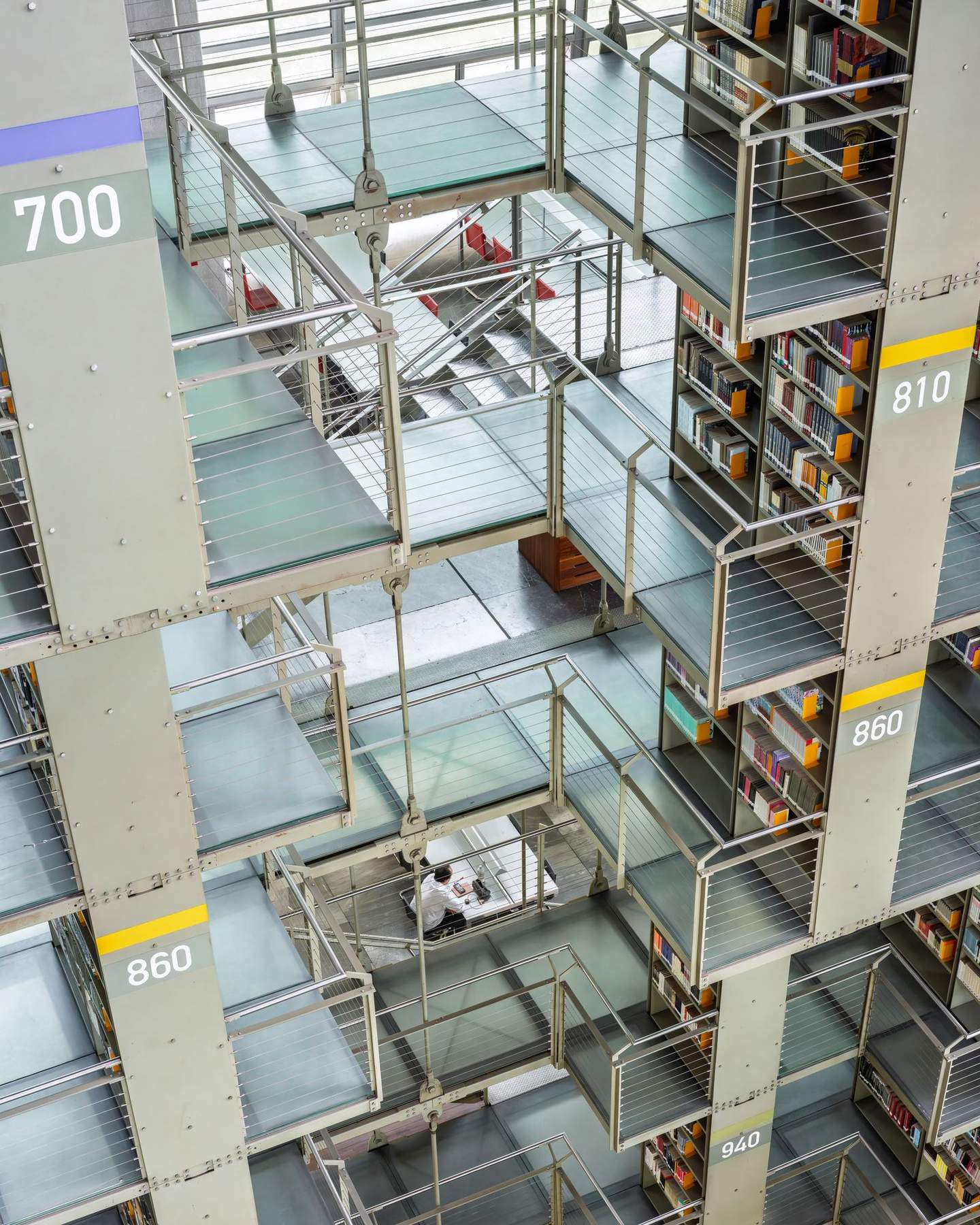 Looking straight down through Biblioteca Vasconcelos suspended stacks, single reader at work table five levels below, cable tensioning hardware and staggered platform edges visible