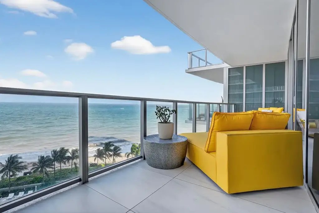 Oceanfront balcony with a yellow sofa, glass railing and palm trees along the shoreline below