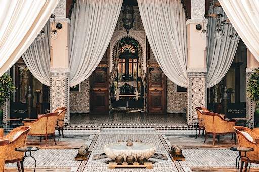 Hospitality interior of a Moroccan riad courtyard with draped curtains, carved doors and a stone fountain